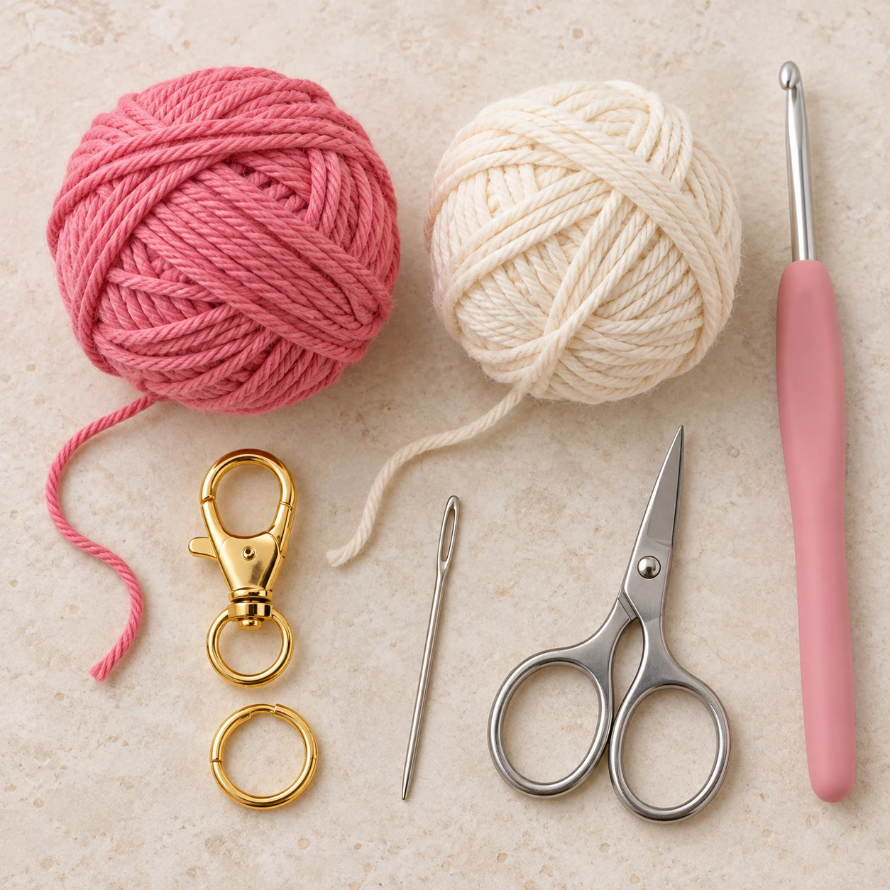 Pink and cream cotton yarn with a crochet hook, yarn needle, scissors, and gold clasp.